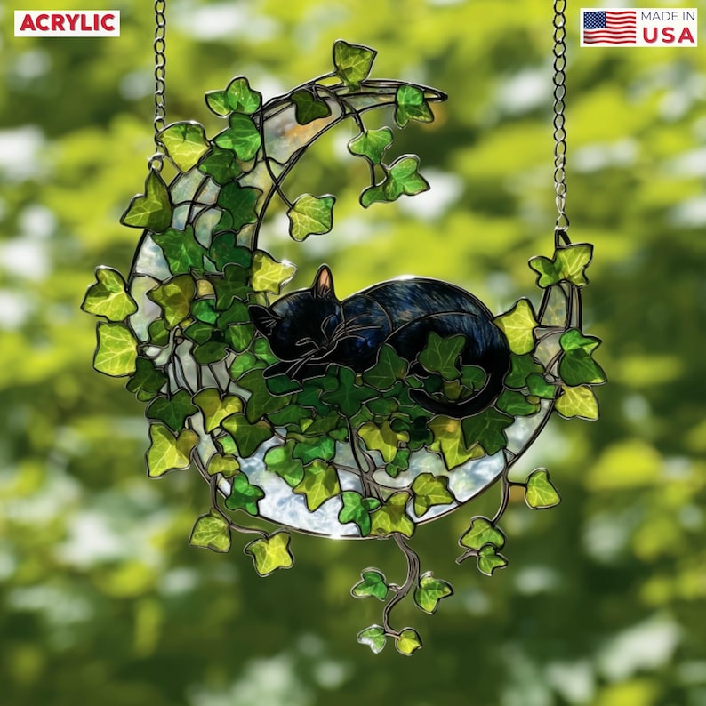May include: Acrylic crescent moon suncatcher with a sleeping black cat nestled in green ivy. The suncatcher is suspended by a chain and features the text "ACRYLIC" and "MADE IN USA".