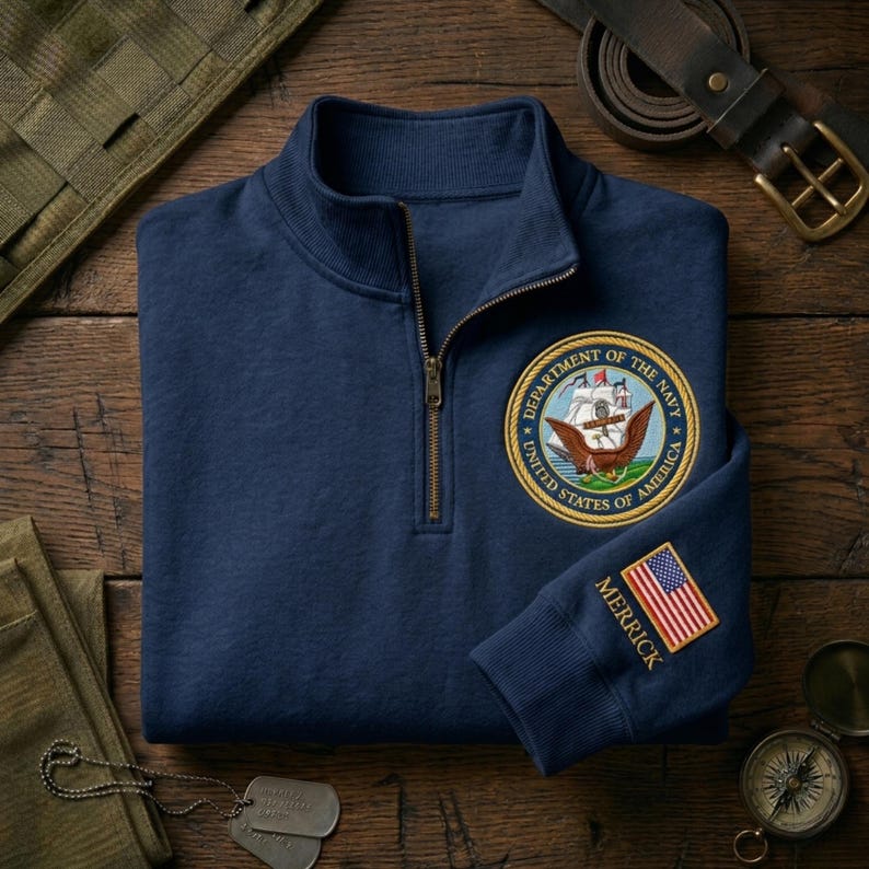 May include: Navy blue quarter-zip sweatshirt with a gold zipper. Features a Department of the Navy patch on the chest and an American flag patch with the name "MERRICK" on the sleeve. The sweatshirt is on a wooden surface.