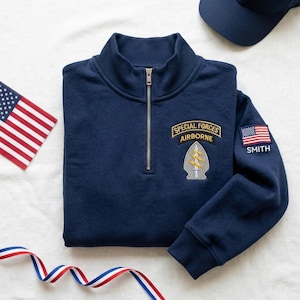 May include: Navy blue sweatshirt with a quarter-zip collar. Features a "Special Forces Airborne" patch and an American flag patch with the name "SMITH". A navy blue cap and an American flag are also visible.