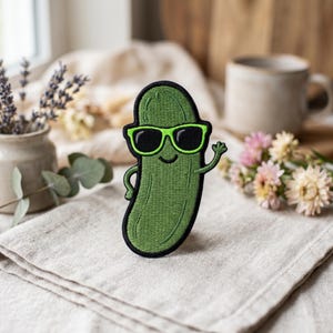 May include: Embroidered patch featuring a smiling pickle wearing sunglasses and waving. The patch is green with black accents and a black border. The design is whimsical and fun, perfect for adding to clothing or accessories.