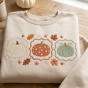 May include: A beige crewneck sweatshirt featuring an embroidered design of three pumpkins in white, orange with white polka dots, and sage green. Each pumpkin is framed by a decorative border, surrounded by embroidered autumn leaves and stars.