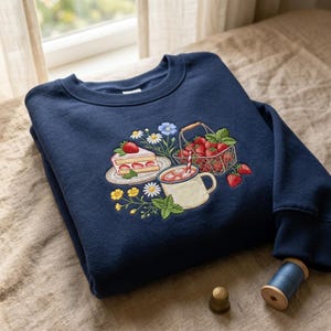 May include: A navy blue crewneck sweatshirt featuring an embroidered design of a slice of strawberry cake on a plate, a basket filled with fresh strawberries, a mug with a red and white striped straw, and various small colorful wildflowers.