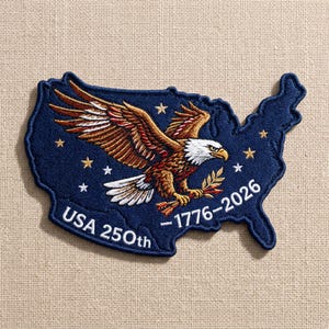 May include: Embroidered patch in the shape of the United States, navy blue with a detailed eagle design. The eagle has brown and white feathers, a white head, and a yellow beak. Text includes "USA 250th" and "1776-2026".