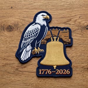 May include: An embroidered patch featuring a white-headed eagle with a yellow beak perched next to a gold-colored bell suspended from a wooden beam. The text 1776-2026 is stitched in gold at the bottom against a dark blue background with a navy border.
