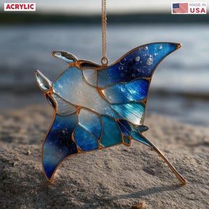 May include: A stained glass-style acrylic manta ray ornament with a gold-colored chain. The ornament features shades of blue and white, with gold-colored outlines. The word "ACRYLIC" is in the upper left corner, and "MADE IN USA" is in the upper right corner.