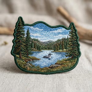May include: Embroidered patch featuring a serene lake scene with a loon swimming in the water. The patch has a green border and depicts trees, mountains, and a blue sky with clouds. The patch is approximately 3 inches wide and 2.5 inches tall.