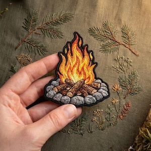 May include: Embroidered patch depicting a campfire with flames in shades of orange and yellow, atop a pile of logs and gray stones. The patch is surrounded by a black border. The background features embroidered greenery.