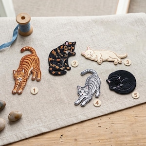 May include: Five embroidered cat patches in various poses and colors. The patches include a tabby cat stretching, a calico cat grooming, a cream-colored cat sleeping, a gray striped cat stretching, and a black cat curled up.