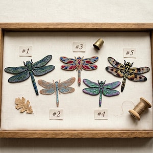 May include: Five embroidered dragonfly patches labeled #1 through #5. Patch #1 is blue and green, #2 is light brown, #3 features red and blue geometric patterns, #4 is purple and green, and #5 is yellow and black. A thimble and spool are included.