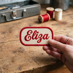 May include: A beige and red embroidered patch with the name "Eliza" in a cursive font. The patch has a red border and is held by a hand. Sewing supplies and spools of thread are in the background.