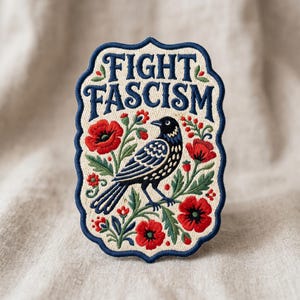 May include: Embroidered patch with a cream background and a navy blue border. The patch features the words "FIGHT FASCISM" in navy blue, a black bird, and red poppies with green leaves. The patch is rectangular with a decorative border.