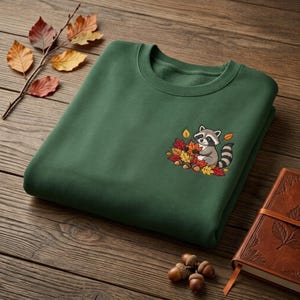 May include: A forest green crewneck sweatshirt featuring an embroidered graphic on the chest of a small raccoon sitting among a pile of colorful autumn leaves and acorns. The design includes shades of orange, yellow, red, and brown.