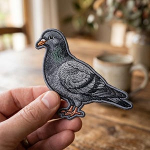 May include: Embroidered patch of a pigeon in shades of gray, with a detailed depiction of feathers and a realistic appearance. The bird has an orange beak and feet, and a green iridescent patch on its neck. The patch is likely for clothing or accessories.