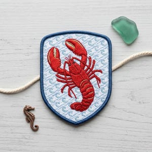 May include: A shield-shaped embroidered patch featuring a bright red lobster centered on a light blue background with a repeating white wave pattern. The patch has a dark blue stitched border and is surrounded by a small copper seahorse charm.