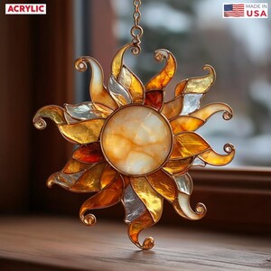 May include: A sun-shaped acrylic ornament with a central, marbled, cream-colored circle and radiating, multi-colored rays. The rays feature gold, silver, and amber hues. The ornament is suspended by a gold chain. The word "ACRYLIC" is in the upper left corner, and "MADE IN USA" is in the upper right corner.
