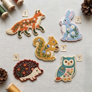 May include: Five embroidered animal patches labeled 1 through 5: a fox with white flowers, a blue rabbit with daisies, a squirrel with blue flowers holding an acorn, a brown hedgehog with orange flowers, and an owl with pink flowers.