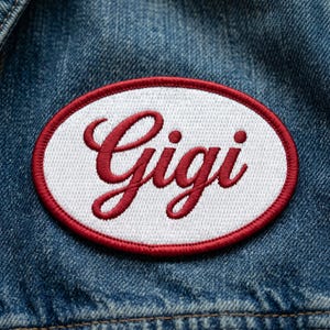 May include: Oval embroidered patch with the name "Gigi" in red script lettering on a white background, framed by a red border. The patch is set against a blue denim fabric.