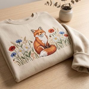 May include: A beige crewneck sweatshirt featuring an embroidered design of a sitting red fox surrounded by colorful wildflowers including red poppies, blue cornflowers, and white daisies with green stems and wheat stalks on the front center.