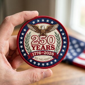 May include: A round patch with a red and blue border featuring white stars. The patch has an eagle with outstretched wings above the text "250 YEARS 1776-2026 WE THE PEOPLE". The patch is held in a hand.