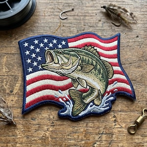 May include: Embroidered patch featuring a largemouth bass leaping from water, set against a waving American flag. The patch is in shades of green, red, white, and blue, with detailed stitching. Fishing-themed.
