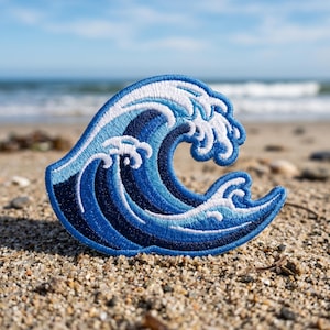 May include: Embroidered patch featuring a stylized ocean wave in shades of blue and white. The wave design has intricate stitching details, with a crest of white foam. The patch is placed on a sandy surface, with the ocean and sky in the background.