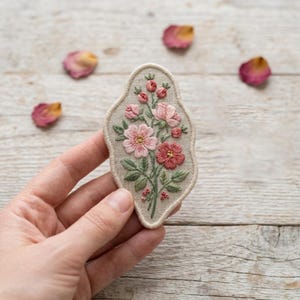 May include: An irregular, shield-shaped fabric patch featuring detailed embroidery of a floral bouquet. The design includes light pink and dark rose-colored flowers, green leaves, and small buds on a neutral beige linen-like background.