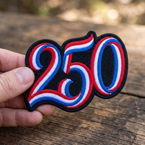 May include: A black embroidered patch with the number "250" in red, white, and blue. The numbers are outlined in white and have a patriotic color scheme. The patch is held in a person's hand.