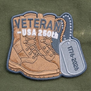 May include: An embroidered patch featuring tan combat boots with the text VETERAN USA 250th in blue and white. A grey dog tag hangs from the boots by a beaded chain, displaying the text 1776-2026. The patch has a blue border and detailed stitching.