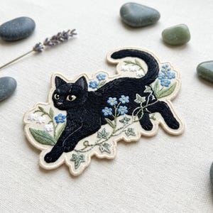 May include: An embroidered patch featuring a black cat in a pouncing position, surrounded by light blue forget-me-not flowers, white lily of the valley, and green ivy leaves on a cream-colored fabric base with a scalloped edge.