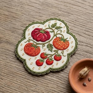 May include: An embroidered fabric patch featuring a cluster of red tomatoes on green vines with leaves. The patch has an off-white background, a scalloped edge with a dark green border, and small yellow floral accents scattered across the surface.