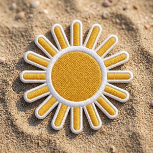 May include: Embroidered sun patch with a yellow center and white and yellow rays. The patch is circular with a textured appearance, set against a sandy background. The design is detailed and bright, suitable for clothing or accessories.
