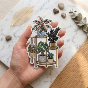 May include: An embroidered patch featuring a gold-toned tiered plant stand holding five potted plants. The plants include a pink and green variegated plant, a dark green leafy plant, a spotted hanging plant, a large-leafed plant, and a small succulent.