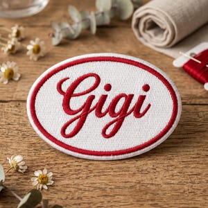 May include: Oval-shaped embroidered patch with the name "Gigi" in red script lettering. The patch has a white background and a red border. The patch is on a wooden surface with flowers and sewing supplies.