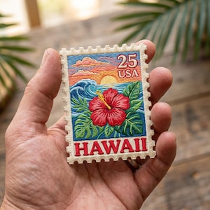 May include: Embroidered patch in the shape of a postage stamp featuring a Hawaiian scene. The design includes a red hibiscus flower, ocean waves, a sunset, and the text "HAWAII" and "25 USA". The patch has a cream-colored border.