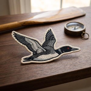 May include: An embroidered patch featuring a loon in flight with wings spread wide. The bird is depicted in shades of black, white, and gray thread with a small red eye. The patch has a cream-colored border and is shaped to follow the bird's outline.
