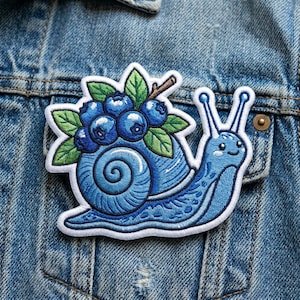May include: A blue embroidered patch featuring a whimsical snail carrying a cluster of blueberries. The snail has a detailed shell and a smiling face. The patch is on a denim jacket.