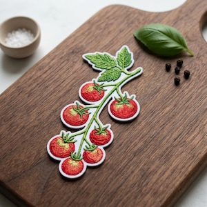 May include: Embroidered patch featuring a cluster of red tomatoes with green leaves on a white background. The patch is placed on a wooden cutting board, alongside a small bowl of salt, a basil leaf, and peppercorns.