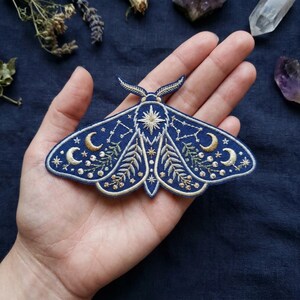 May include: Embroidered navy blue moth patch featuring celestial designs. The patch has a central star, crescent moons, constellations, and floral accents in gold and white thread. The moth's wings are detailed with intricate stitching.
