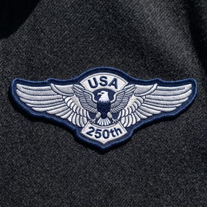 May include: Embroidered patch featuring a white eagle with outstretched wings, the word "USA" above, and "250th" below. The patch has a blue border and is set against a dark gray fabric background.