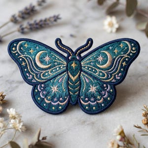 May include: Embroidered patch in the shape of a moth with teal wings and a navy blue border. The wings feature crescent moons, stars, and decorative swirls in gold, pink, and cream thread. The moth's body is teal and gold.