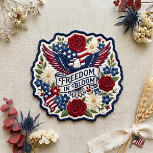 May include: Embroidered patch with a patriotic theme. Features an eagle with outstretched wings, the American flag, and the text "FREEDOM IN BLOOM." The design includes red roses, blue flowers, and white daisies, all set against a cream background.
