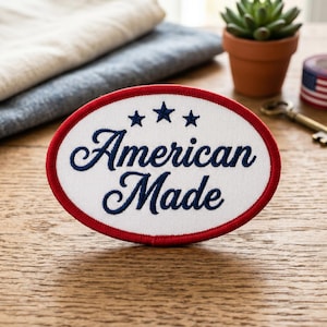 May include: Oval patch with a white background, red border, and the words "American Made" in navy blue script. Three navy blue stars are above the text. The patch is on a wooden surface.