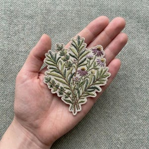 May include: An embroidered patch featuring a botanical arrangement of green leaves, stems, and small purple flowers on a cream-colored fabric base. The design displays detailed needlework with various shades of green and purple thread throughout.