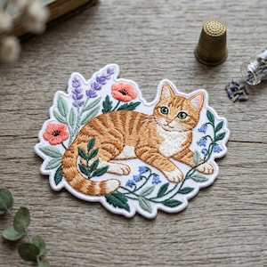 May include: An embroidered patch featuring an orange tabby cat lying down surrounded by lavender, pink poppy flowers, and small blue bell-shaped flowers. The cat has white markings on its chest and paws, with green eyes and a detailed striped coat.