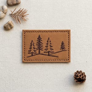May include: A rectangular tan fabric patch featuring a dark brown embroidered design of four evergreen trees on a rolling landscape. The patch has a stitched border around the entire perimeter, creating a clean and defined edge for the rectangular shape.