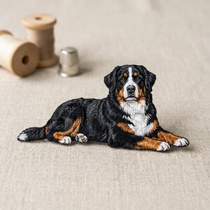 May include: Embroidered Bernese Mountain Dog patch, featuring black, white, and brown fur. The dog is lying down, with its paws extended. Two wooden spools and a silver thimble are in the background.
