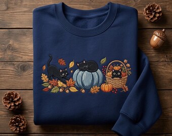 Könnte beinhalten: Ein marineblaues Sweatshirt mit Rundhalsausschnitt und einer Stickerei von drei schwarzen Katzen, Herbstblättern, Eicheln, Kürbissen und einem geflochtenen Korb. Eine Katze streckt sich, eine schläft auf einem blauen Kürbis und eine sitzt im Korb.