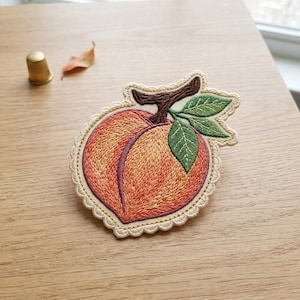 May include: An embroidered patch shaped like a peach with a scalloped cream-colored border. The fruit features shades of orange and pink thread, a dark brown stem, and two green leaves. The design is detailed with visible stitching throughout.