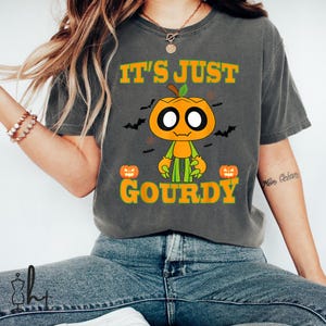 Op de afbeelding: Donkergrijs T-shirt met de tekst "IT'S JUST GOURDY" in oranje. Het shirt heeft een cartoon pompoen karakter met een groene stengel en armen, en twee jack-o'-lanterns. Zwarte vleermuizen zijn ook in het ontwerp opgenomen.
