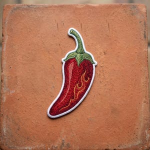 May include: An embroidered patch shaped like a chili pepper. The pepper is deep red with a green stem and features yellow flame-like embroidery details along the side. The entire patch is outlined with a thick white border.